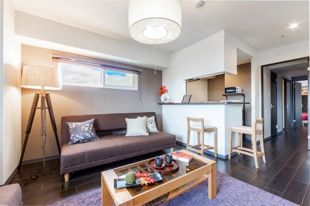 A modern living room with a sofa, coffee table, and a lamp. There is a kitchen area with a microwave and a refrigerator.