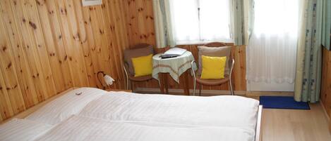 Una habitación con paneles de madera, una cama, una pequeña mesa con sillas y una ventana con cortinas.