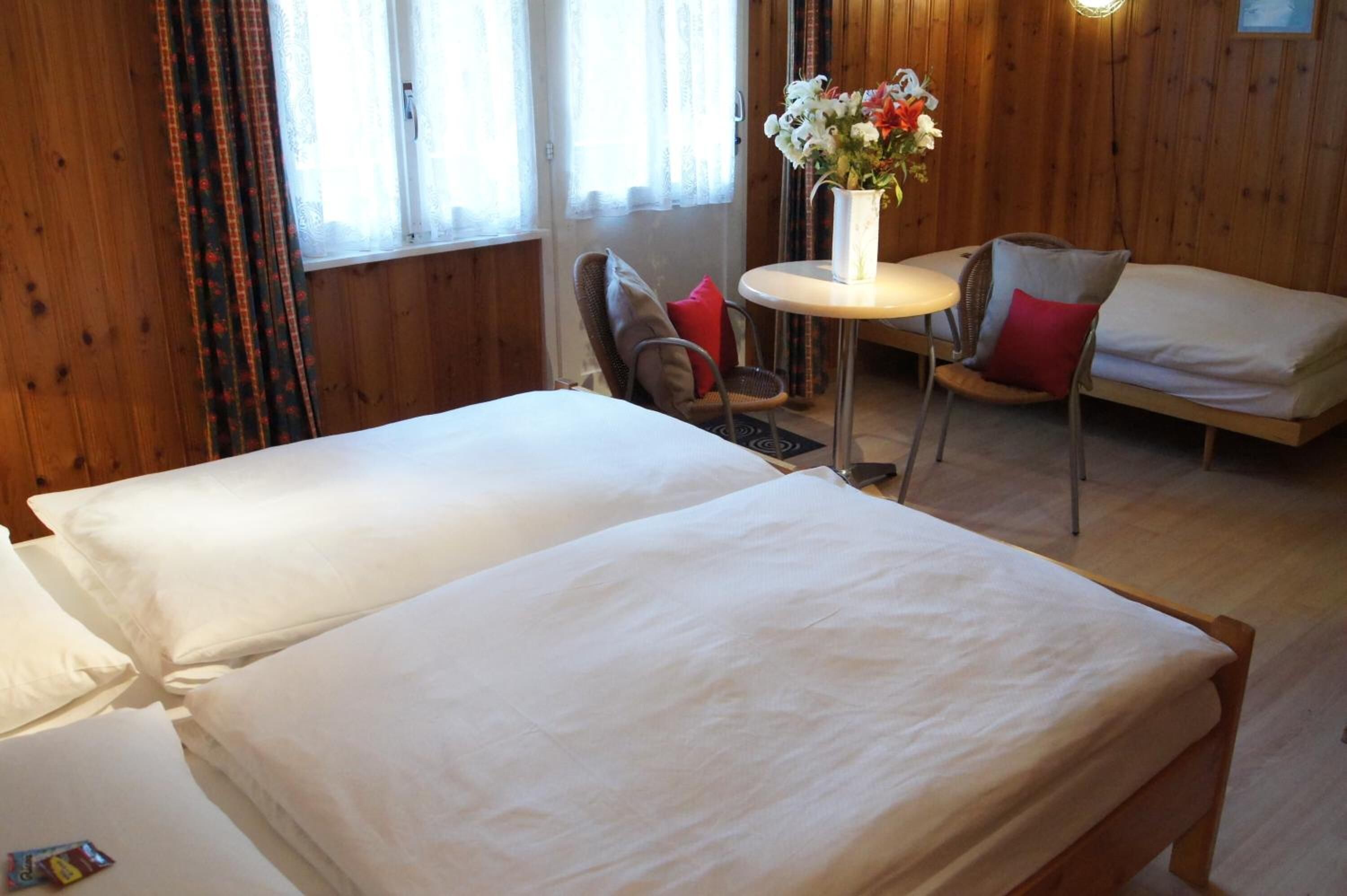 Una habitación con dos camas, una mesita con un jarrón de flores y una silla con un cojín rojo.