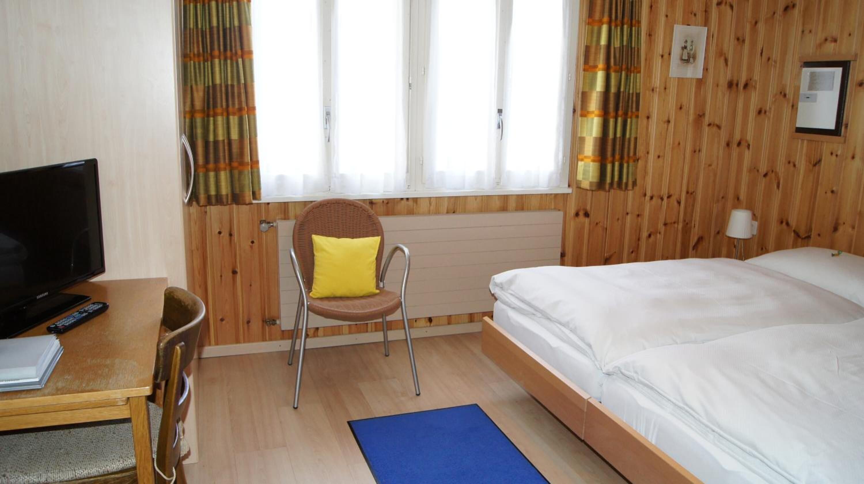 A room with a bed, a chair, a desk with a TV, and a window with curtains.