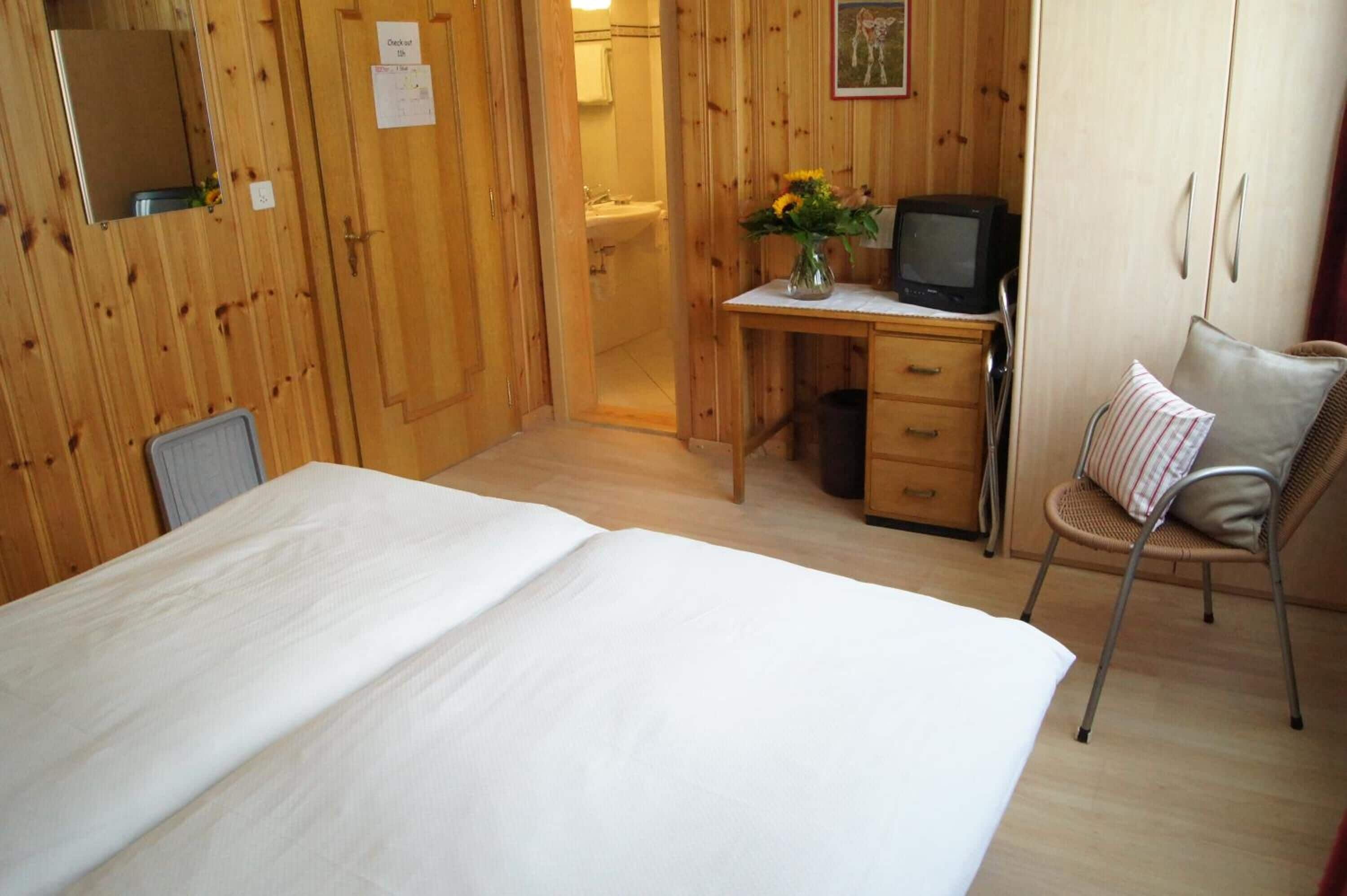 Ein Zimmer mit Holzwänden, einem Bett, einem Schreibtisch mit Fernseher und einem Sessel mit Kissen.
