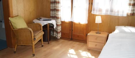 Una habitación con una cama, una silla, una mesita con un ordenador portátil, una lámpara y cortinas.