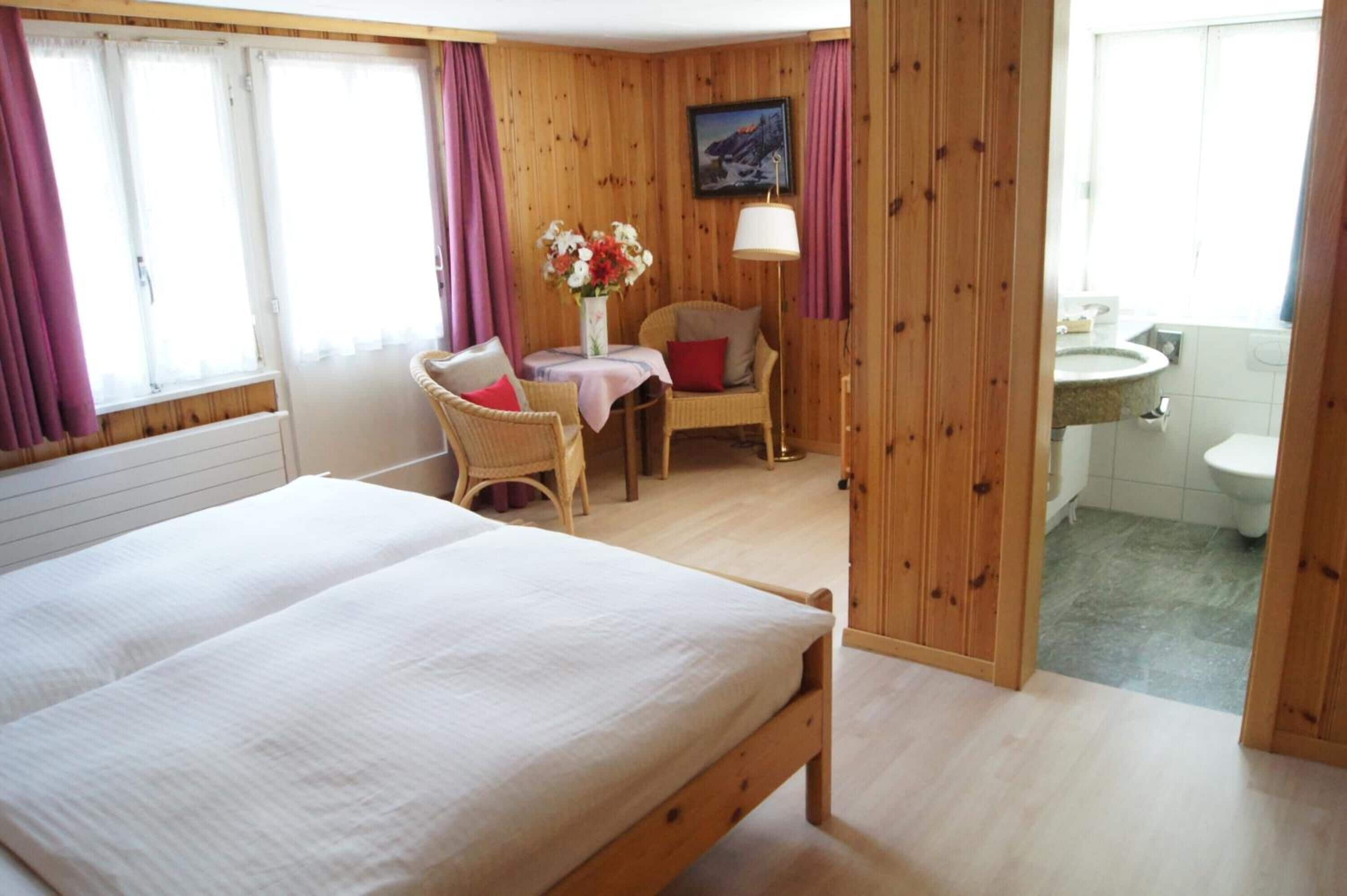 Un dormitorio con una cama grande, una mesita con un jarrón de flores, una silla y vistas al baño.