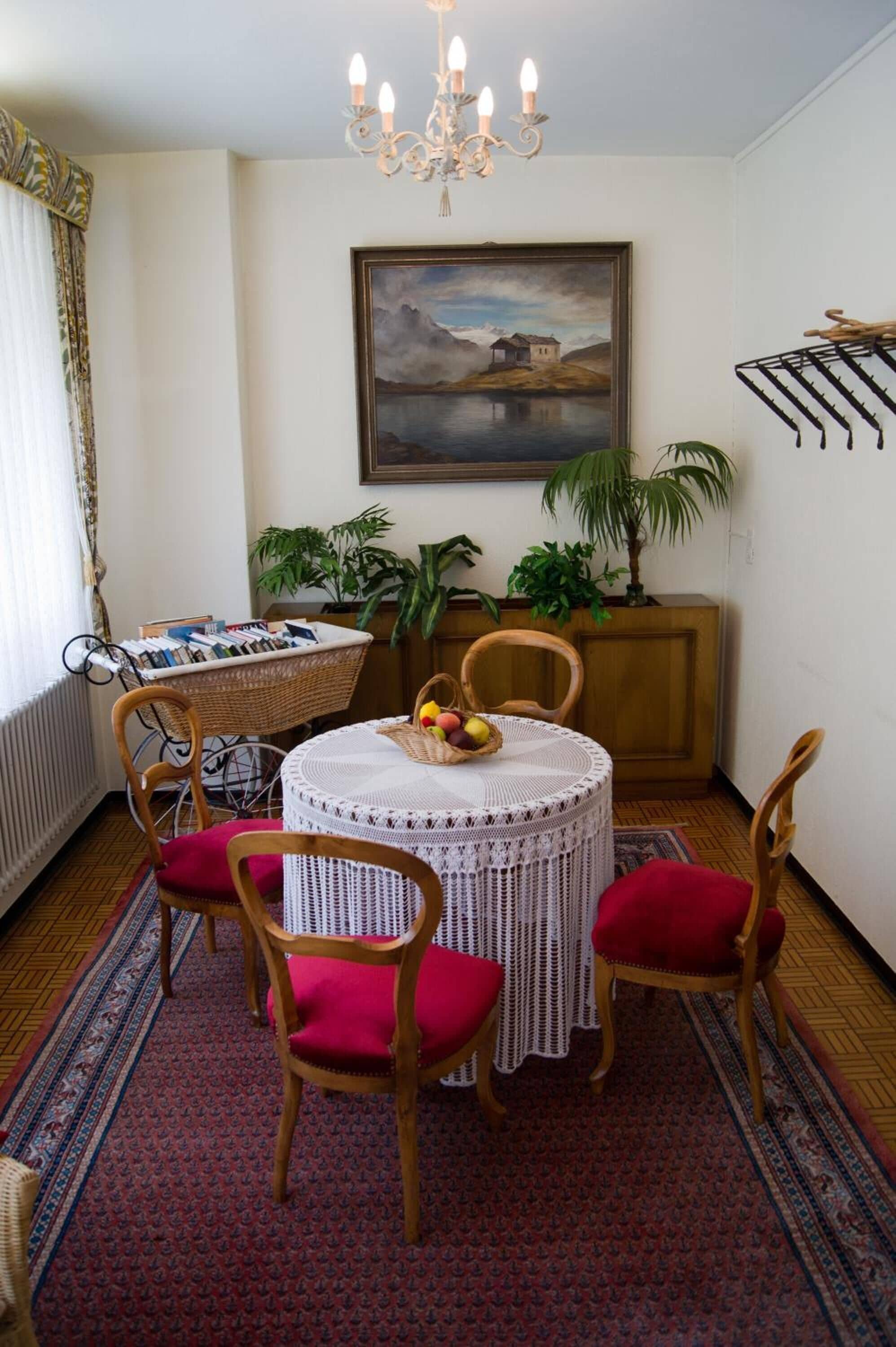 A dining area with a round table, four chairs, a chandelier, a painting, and potted plants.