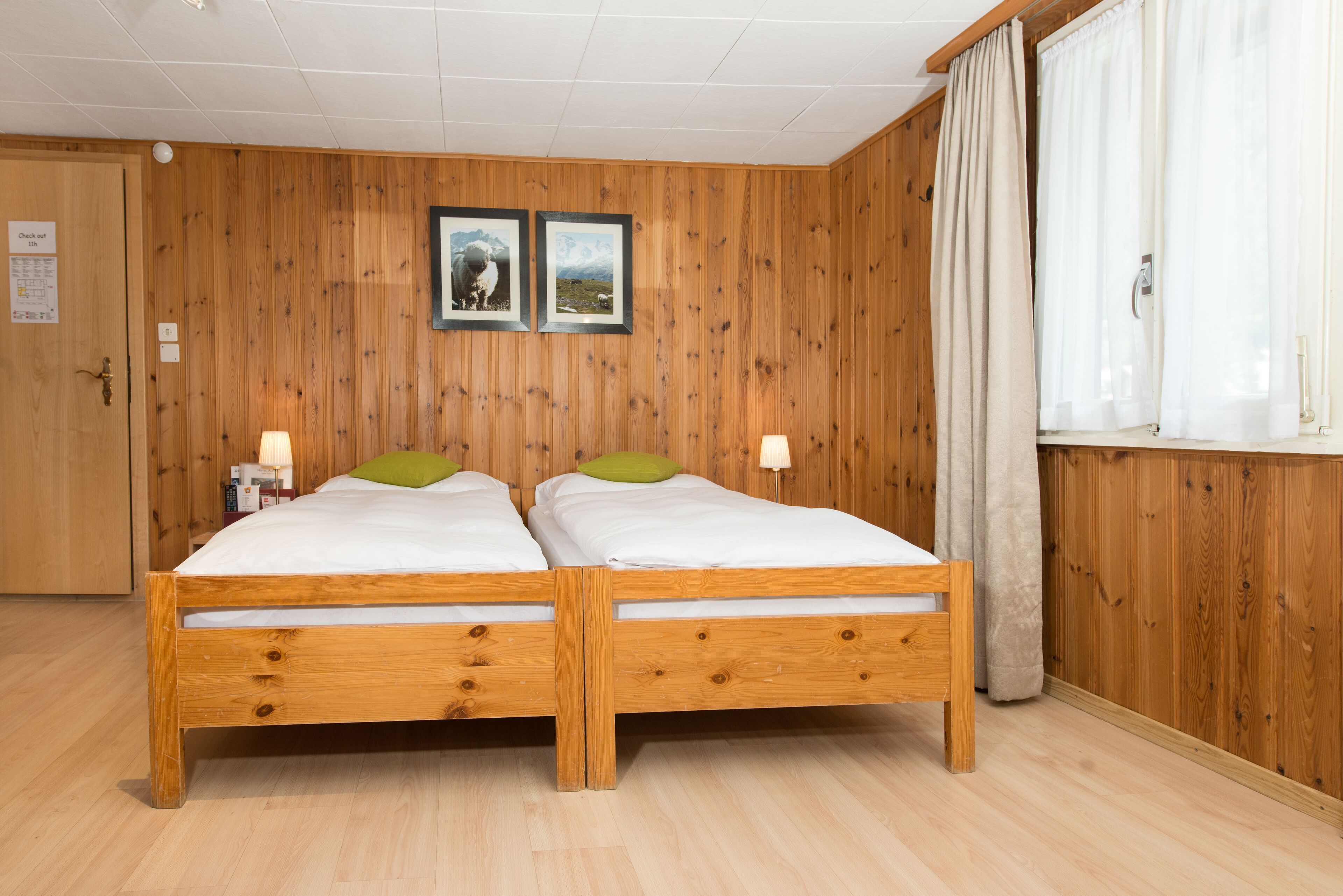 A double wooden bed with two pillows, a wooden floor, a wooden wall with two framed pictures, and a window with curtains.