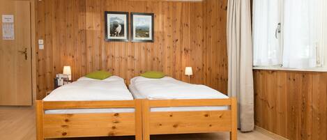Een tweepersoons houten bed met twee kussens, een houten vloer, een houten wand met twee ingelijste foto's en een raam met gordijnen.