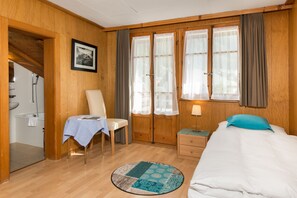 A hotel room with a bed, a chair, a table with a tablecloth, a small bedside table with a lamp, a window with curtains, and a picture on the wall.