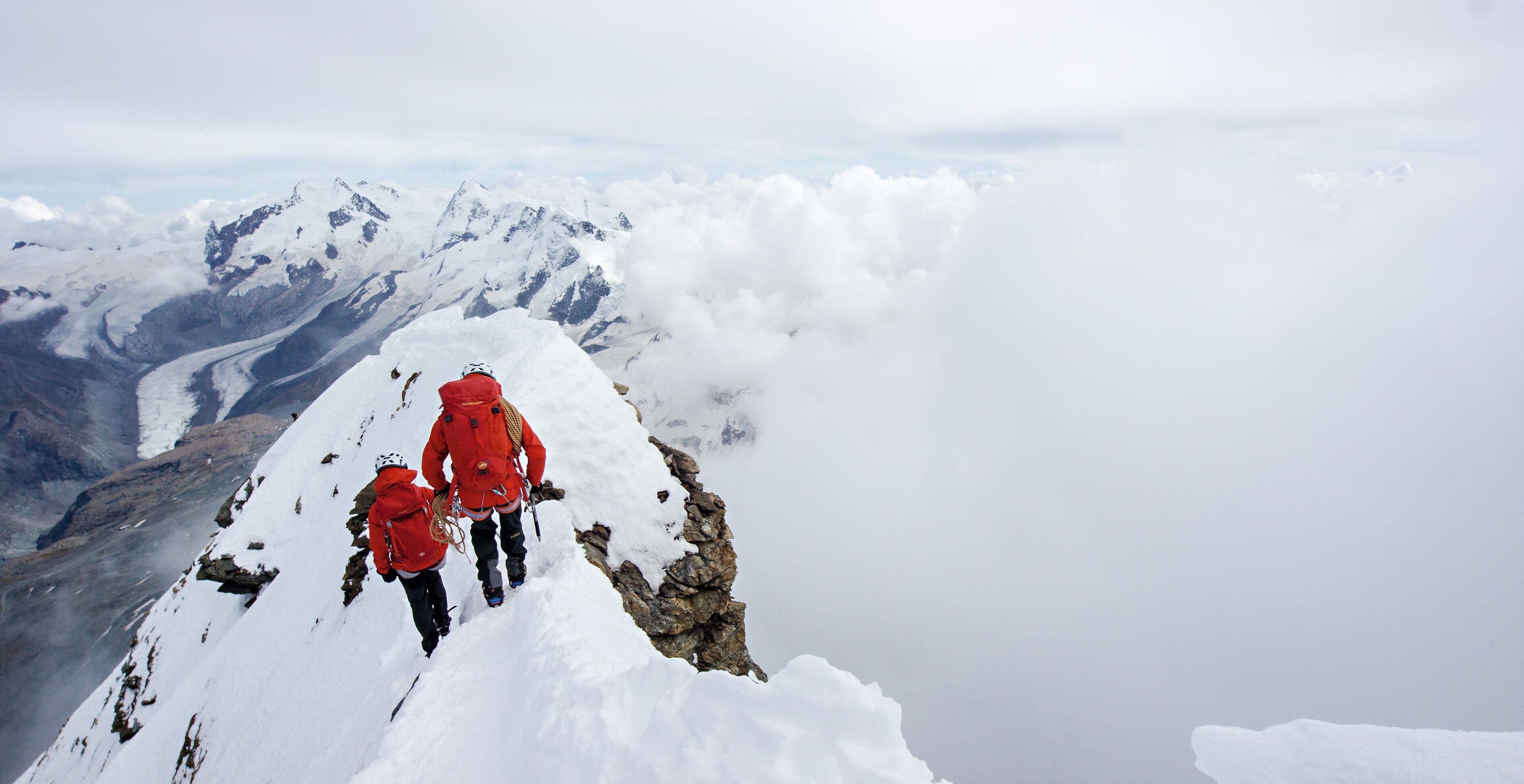 Un pico nevado de montaña con dos excursionistas con chaquetas rojas.