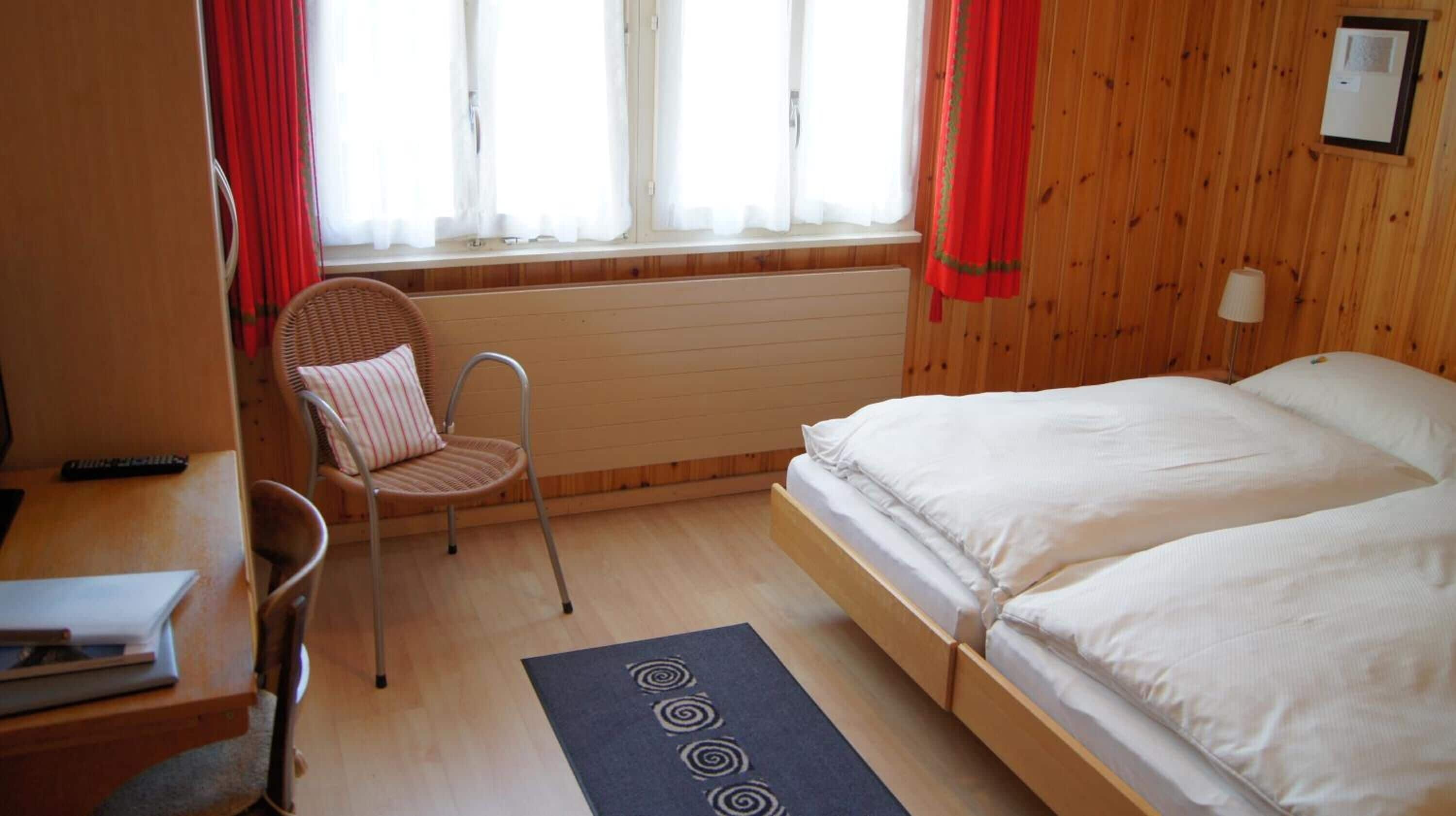 Una habitación pequeña y acogedora con una cama de madera, una silla, un escritorio y una ventana con cortinas.