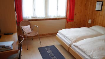 A small, cozy room with a wooden bed, a chair, a desk, and a window with curtains.