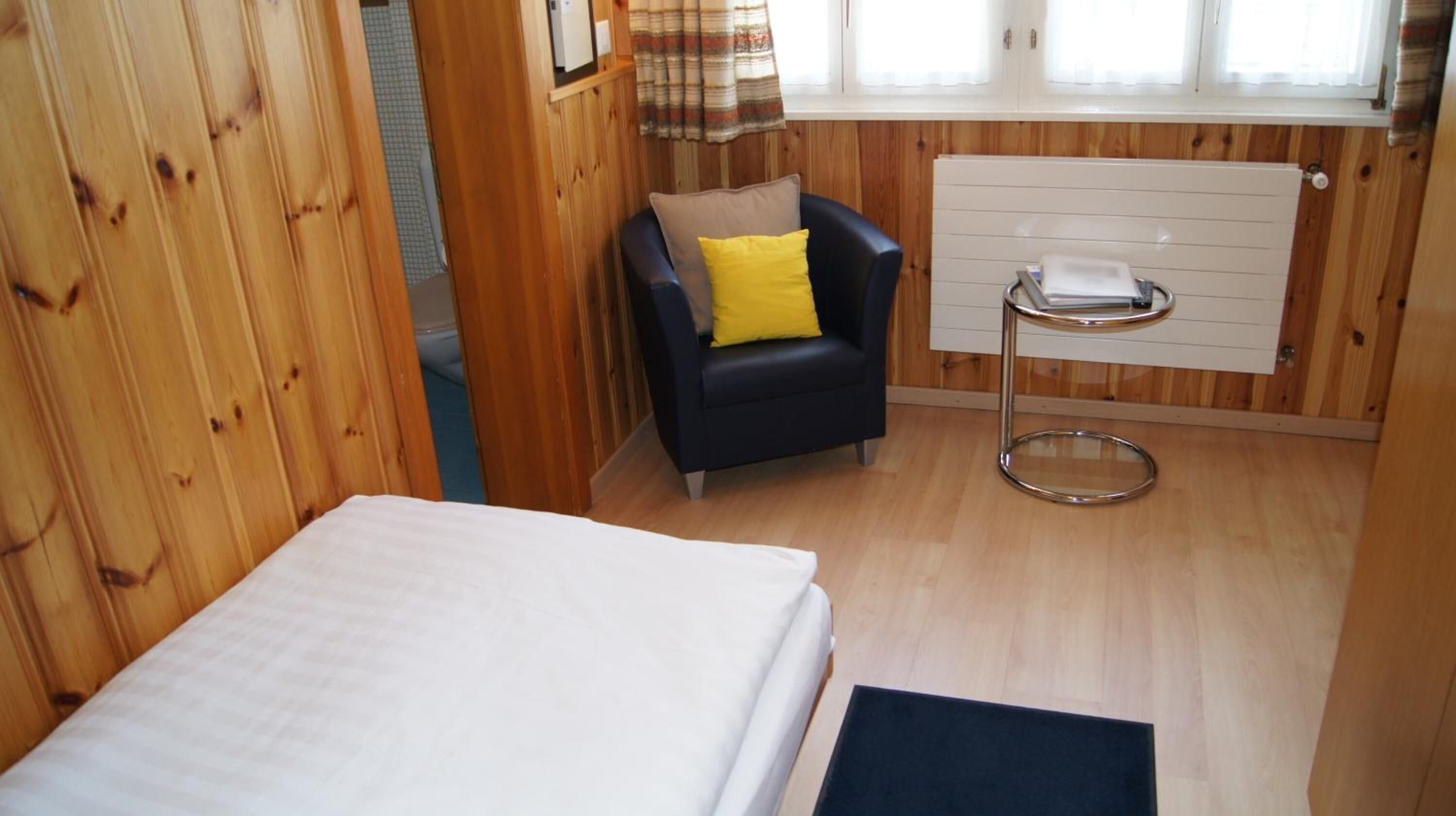 Una habitación pequeña y acogedora con paredes de madera, una cama individual, una silla con un cojín amarillo y un radiador.