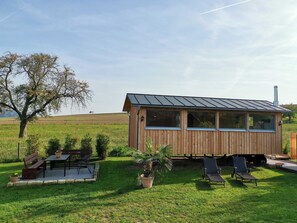 Eine Holzhütte mit einem Metall-Dach, einer Veranda mit Stühlen und Tischen und einem grünen Feld im Hintergrund.