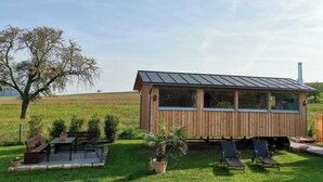 A wooden cabin with a metal roof, a deck with chairs and tables, and a grassy field in the background.