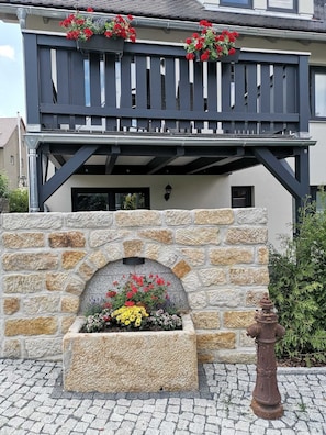 A building with a stone facade, a balcony with red flowers, and a fire hydrant. - Apartment 2-6 Personen DG - Apartmenthaus Vetter (Gohrisch)