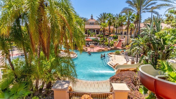 A resort pool area with palm trees, a central pool, and a building with a red roof in the background.