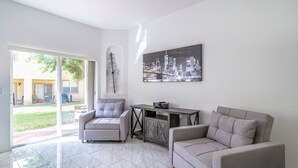 A spacious living area with a large window, a grey sofa, a grey armchair, a small table, and a cityscape painting on the wall.