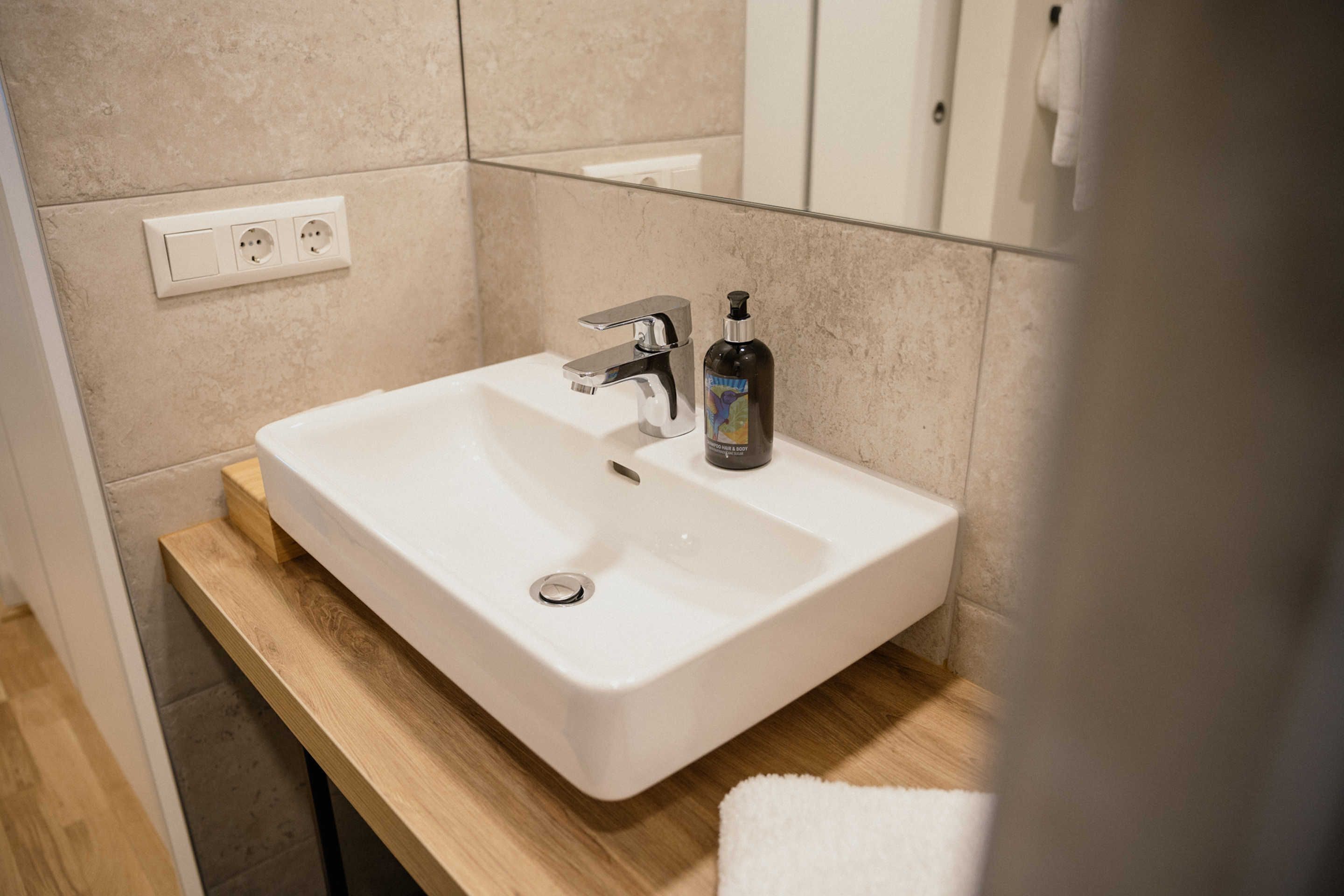 A bathroom with a white sink, a wooden vanity, a mirror, and a bottle of hand soap.