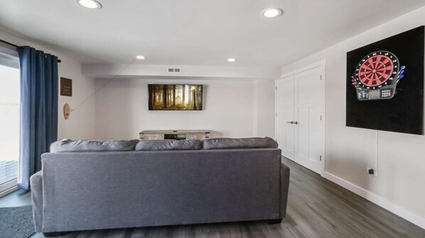 A living room with a grey sofa, a painting of a forest, a dartboard, and a white door.