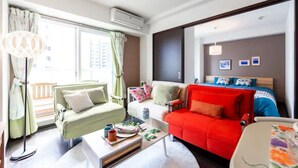 Habitació d'hotel moderna amb un llit gran, un sofà verd, un sofà vermell i una taula de cafè blanca. Hi ha una finestra amb cortines i una làmpada de peu.