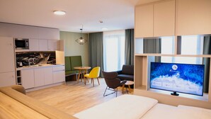 Moderni hotellihuone, jossa on taulutelevisio, sohva, ruokailutila pöytineen ja tuoleineen sekä pieni keittiönurkkaus.