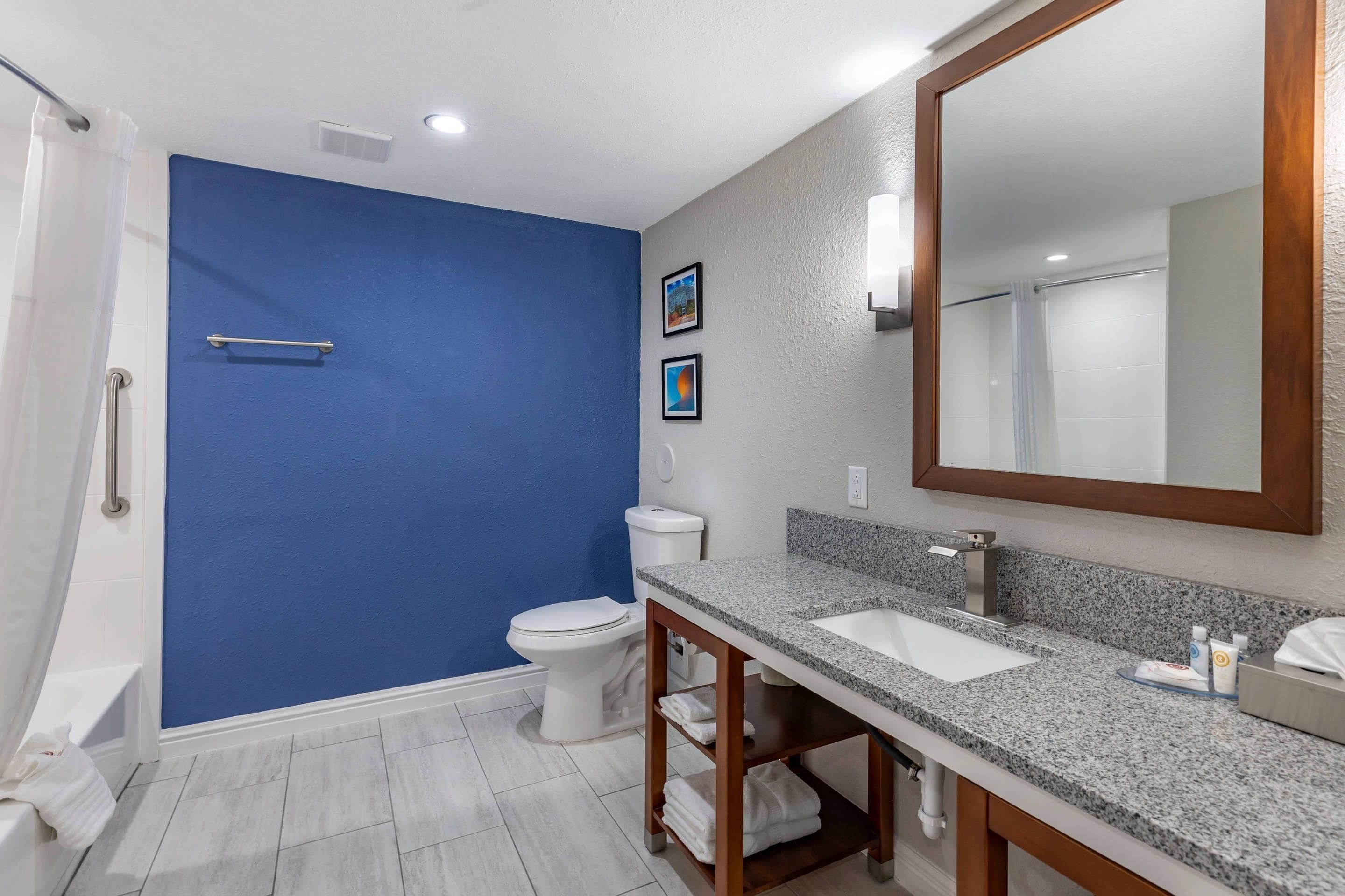 Une salle de bain avec un mur d’accent bleu, des toilettes blanches, un lavabo avec un comptoir en granit et un grand miroir.
