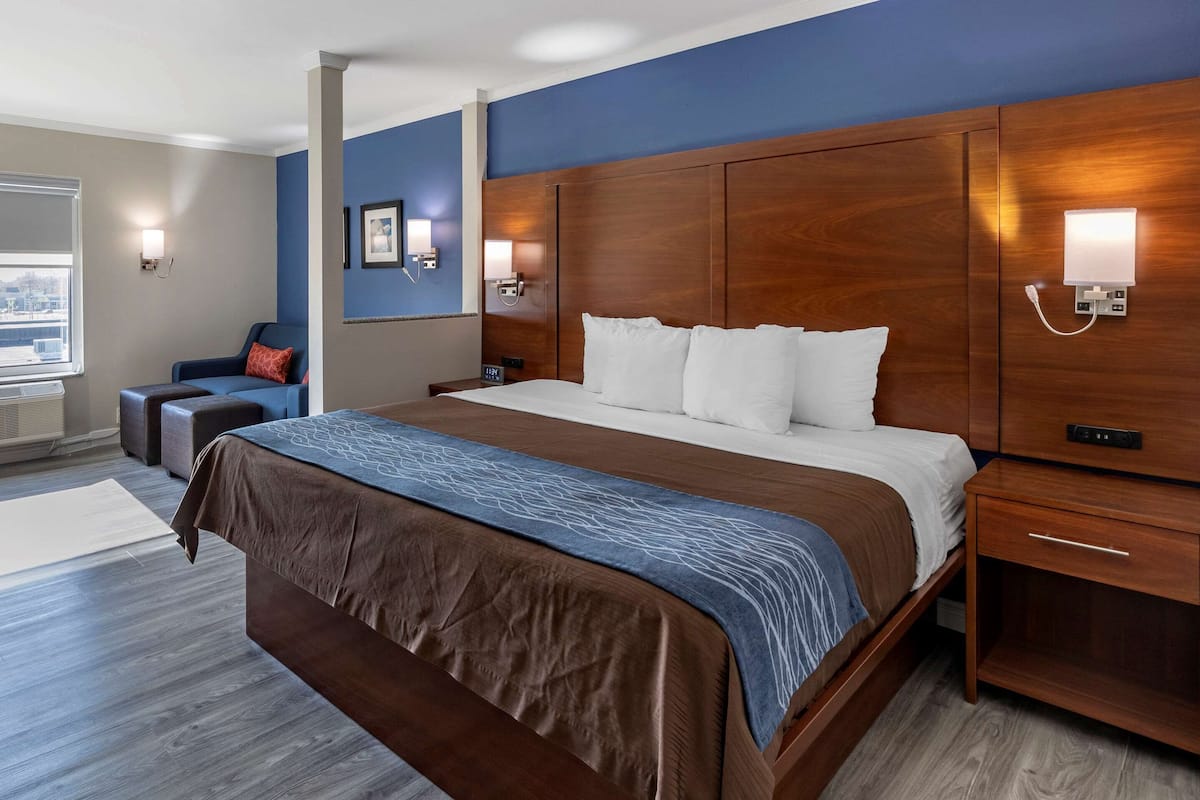 A hotel room with a large bed, a wooden headboard, bedside tables, and a seating area with a chair and ottoman.