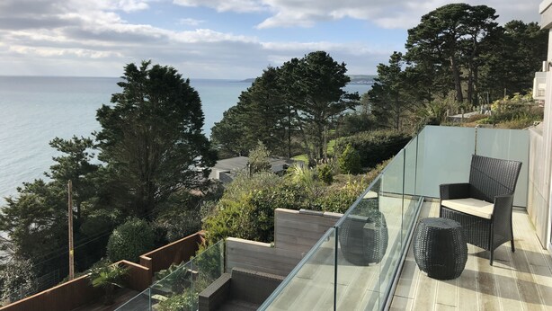 En balkon med gelænder af glas, der ud til et kystlandskab, med en fletstol og et rundt bord.