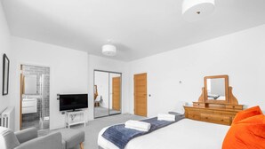 A modern hotel room with a large bed, a television, a wooden dresser, and a bathroom visible through a glass door. - Panmure Apartment - golf course views (Monifieth)