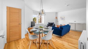 A modern living room with a glass dining table, blue sofa, and a kitchen area with white cabinets. - Panmure Apartment - golf course views (Monifieth)