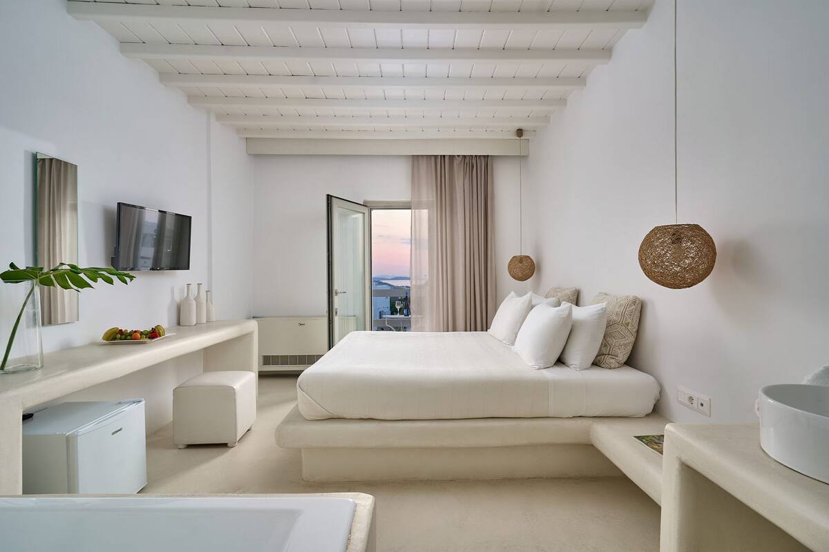 A modern bedroom with a large bed, a small desk, a TV, and a view of the outdoors.