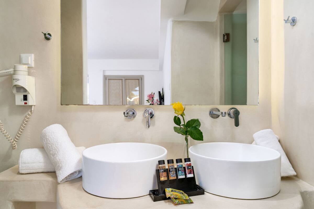 A bathroom with two white sinks, a mirror, a flower vase, and toiletries.