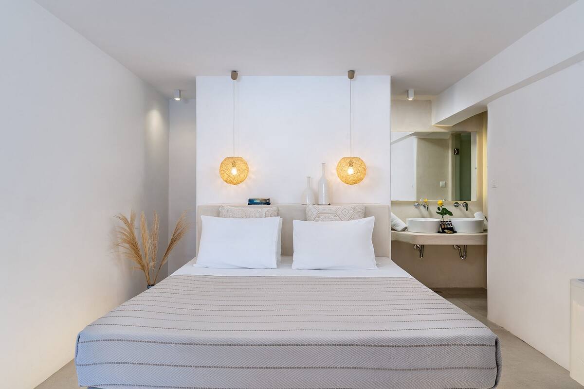 A modern bedroom with a large bed, two bedside tables, a nightstand, a bedside lamp, and a bathroom with a sink and mirror.