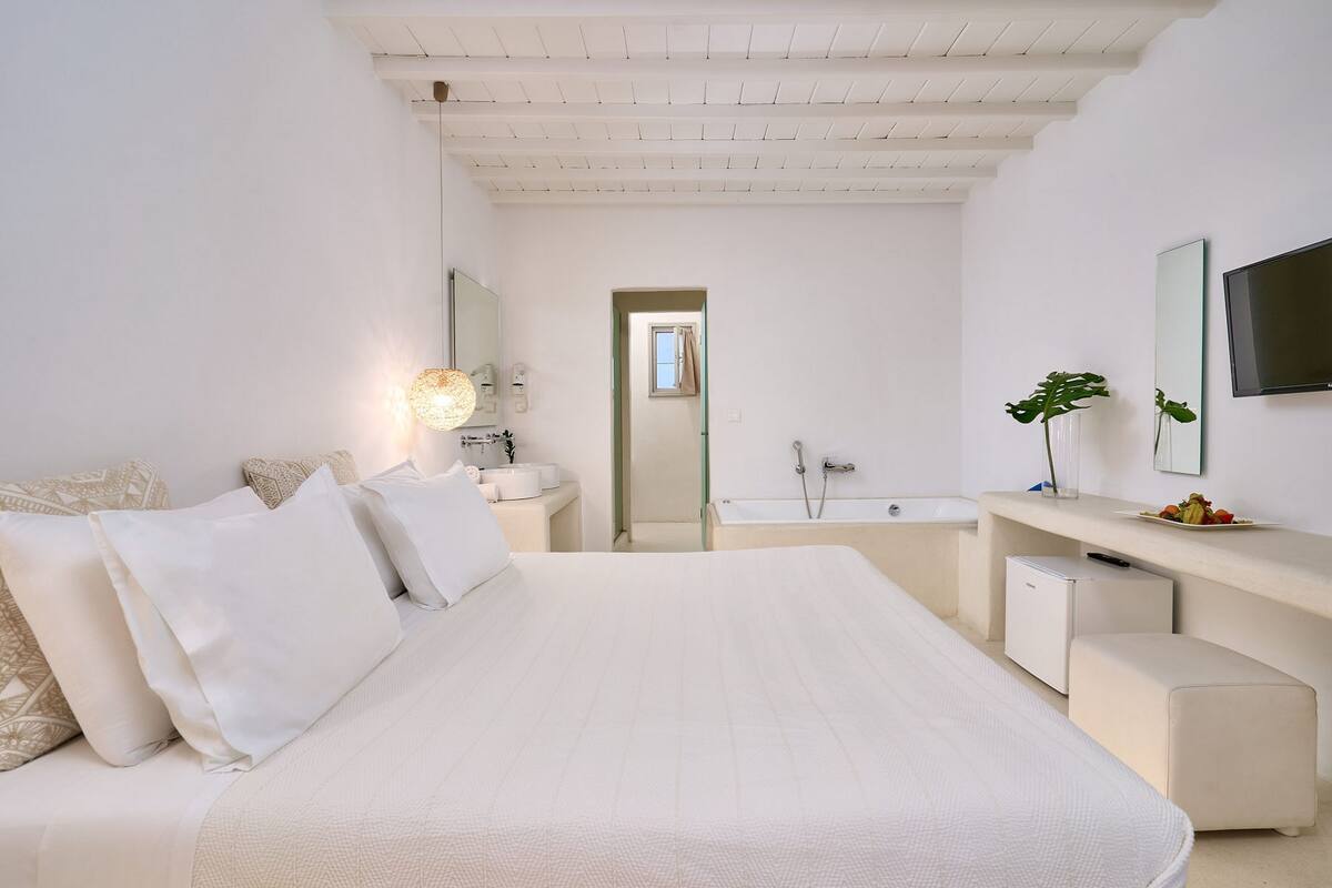 A modern bedroom with a large bed, a bedside table with a lamp, a mirror, and a small refrigerator.