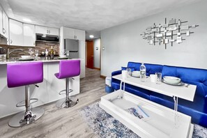 A modern kitchen with white cabinets, a stainless steel refrigerator, and a microwave. There is a dining area with a blue sofa and a white table.