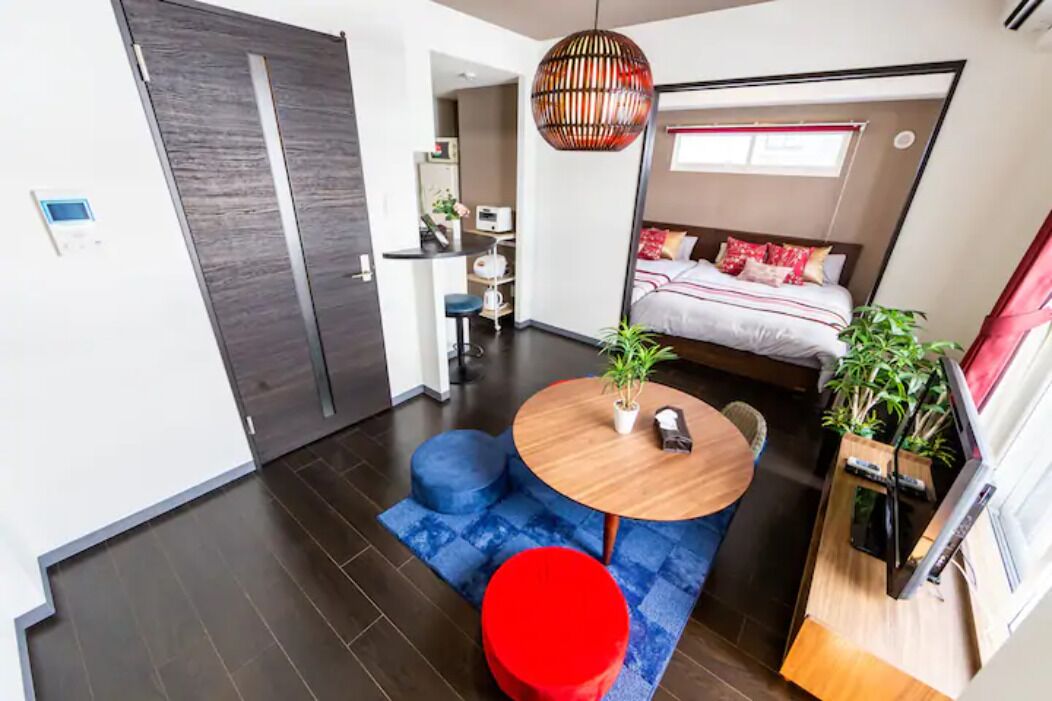A compact living space with a bed, a dining area with a round table and two stools, a small kitchenette, and a large window providing natural light.