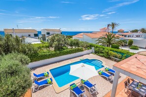 A swimming pool with lounge chairs and an umbrella, surrounded by greenery and buildings with red-tiled roofs. - Menorca Velasco (Cala´n Blanes)