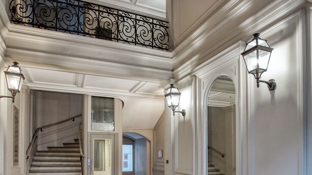 Un vaste hall avec un sol en marbre, une rampe en fer forgé décorée et des luminaires fixés aux murs.