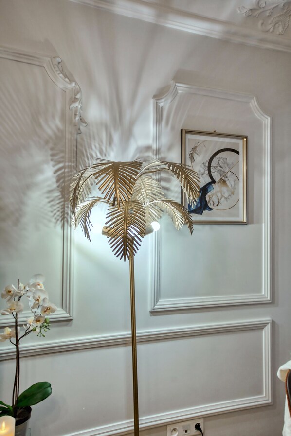 Une pièce avec une lampe de sol décorative dotée d’un pied doré et d’un abat-jour aux motifs évoquant des plumes, une œuvre d’art encadrée accrochée au mur et une plante orchidée en pot.