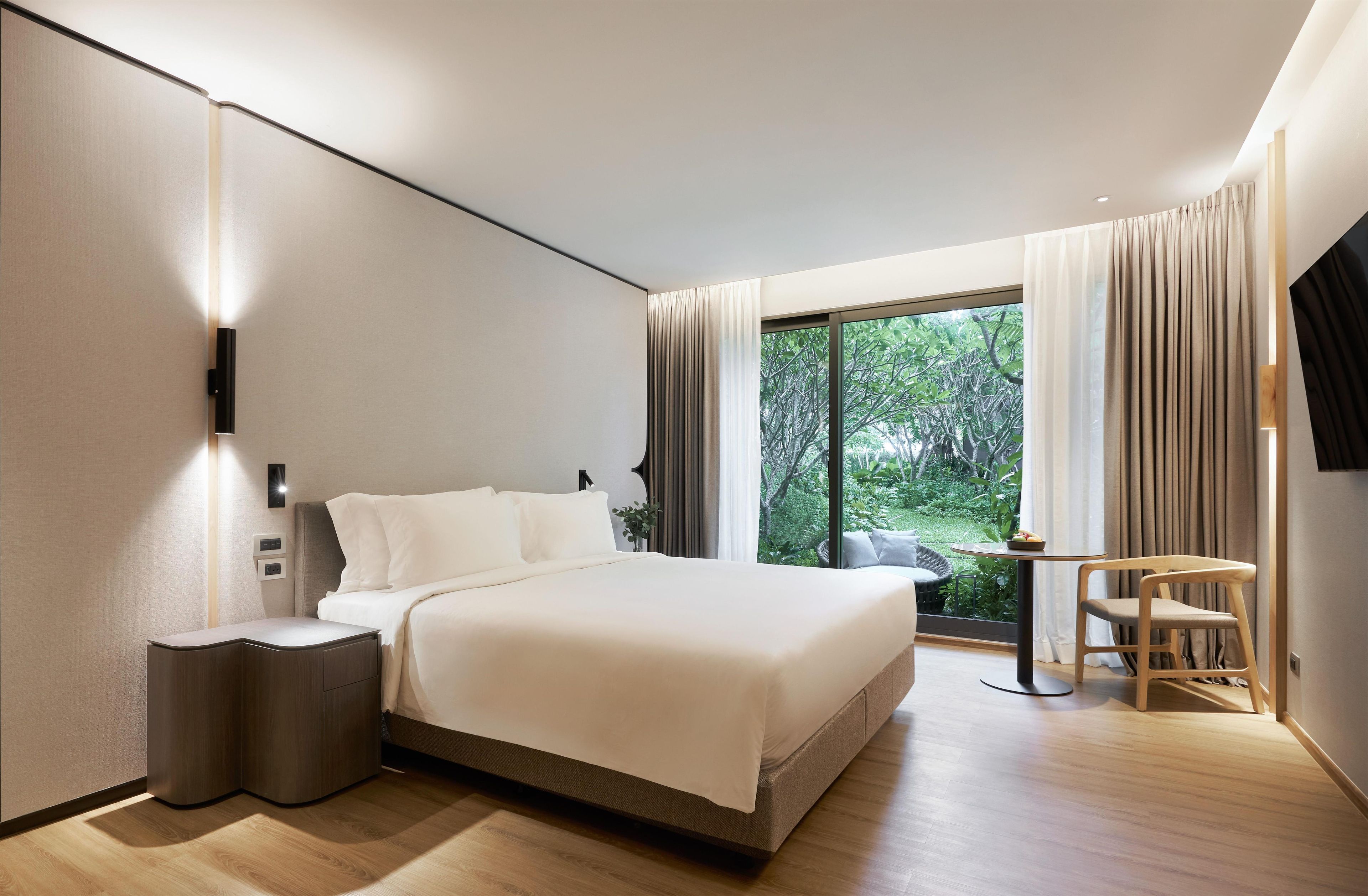 Une chambre d’hôtel moderne avec un grand lit, des tables de chevet, une petite table avec une chaise et une vue sur la verdure par la fenêtre.