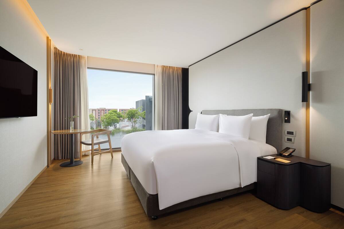 A hotel room with a large bed, a bedside table with a phone, a small table with a chair, and a TV mounted on the wall.