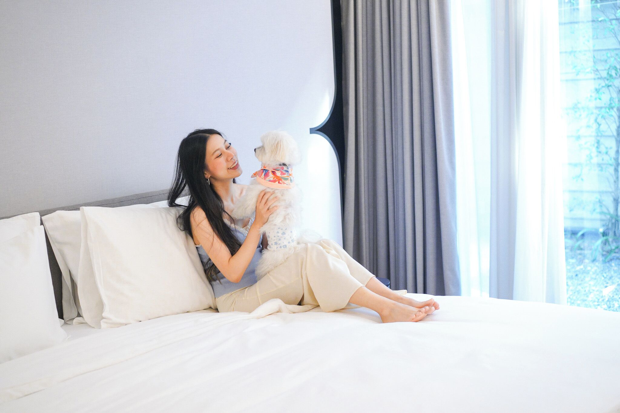 A person sitting on a bed with a dog, in a room with a large window and curtains.