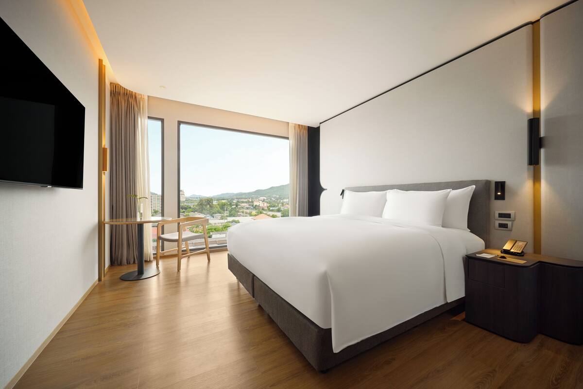 A modern hotel room with a large bed, a flat-screen TV, a bedside table with a lamp, and a view of the city.