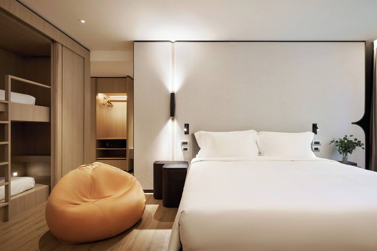 A modern hotel room with a large bed, a bean bag chair, a bedside table, and a wardrobe.