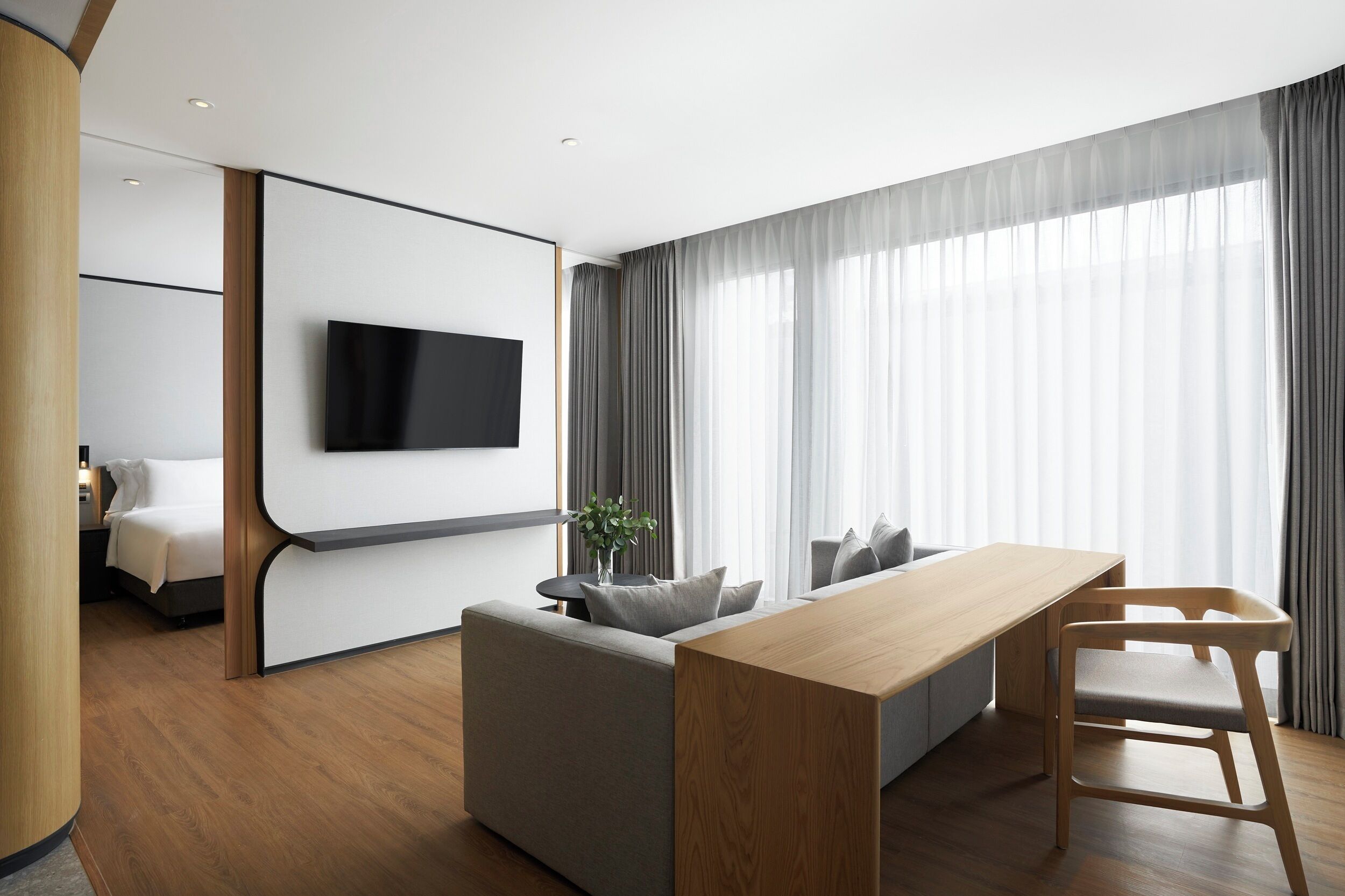 Una habitación de hotel moderna con piso de madera, un sofá gris, una mesa de comedor de madera y un televisor montado en una pared blanca.