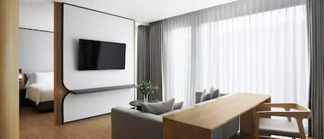 Una habitación de hotel moderna con piso de madera, un sofá gris, una mesa de comedor de madera y un televisor montado en una pared blanca.