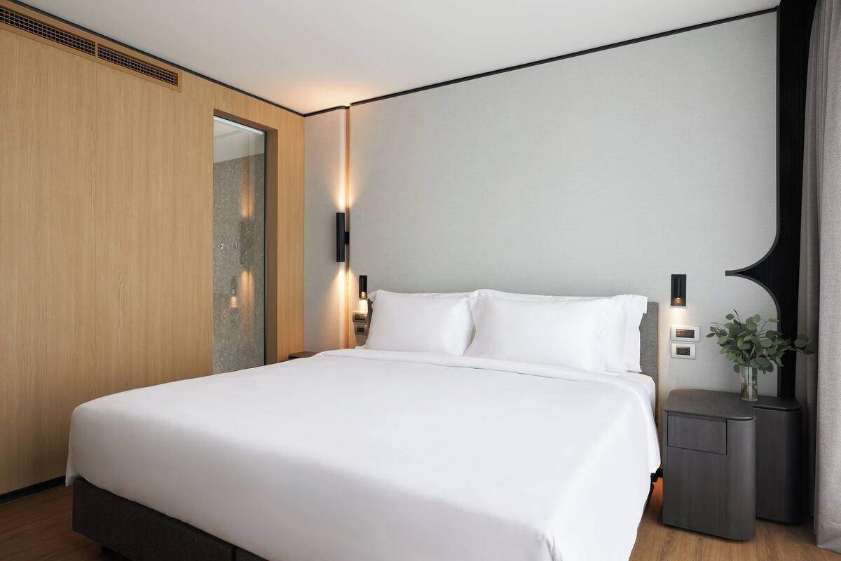 A modern hotel room with a large bed, a wooden wardrobe, a mirror, and a small bedside table with a vase.