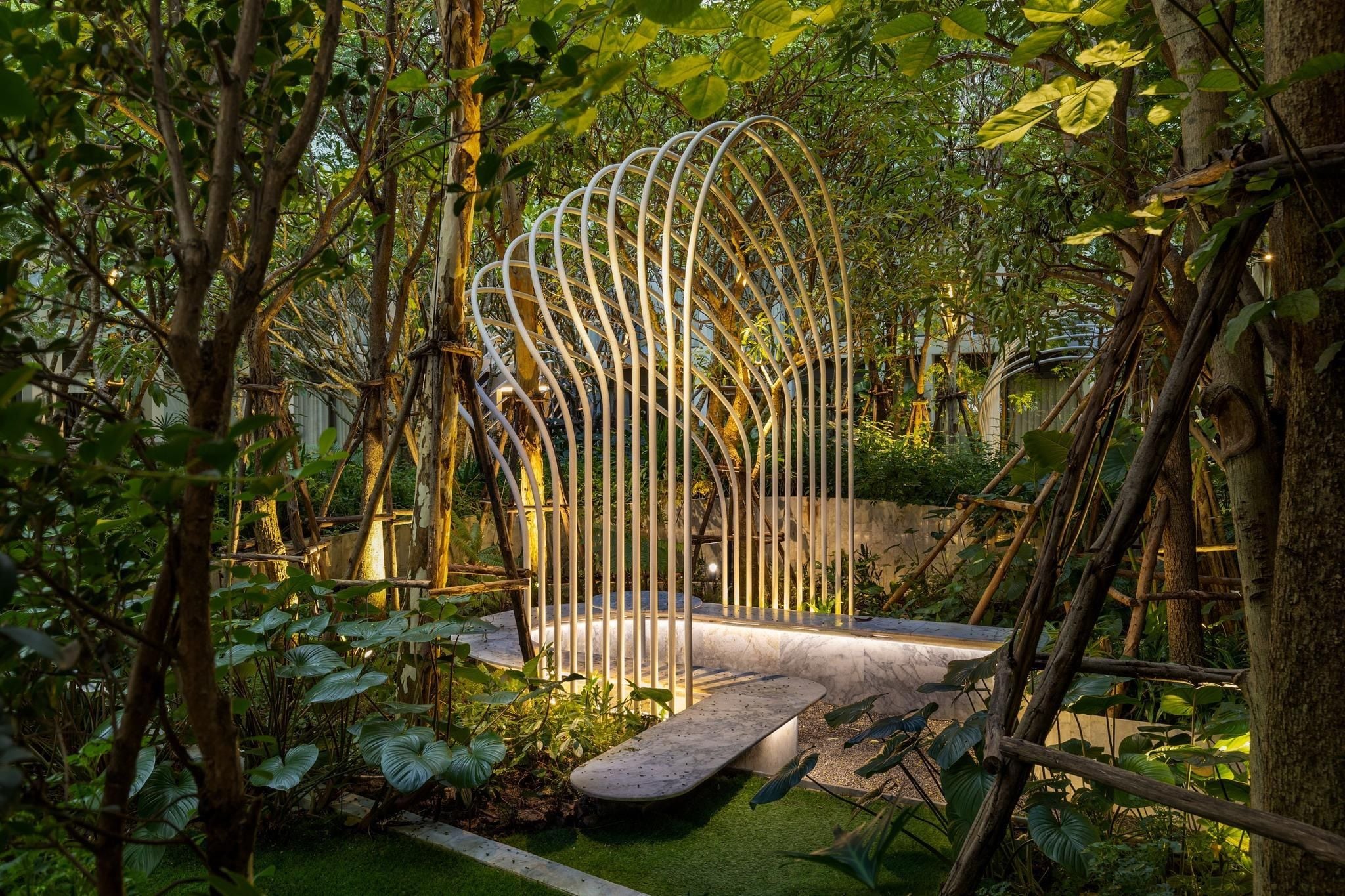 Een tuin met een metalen sculptuur, een betonnen bank en weelderige begroeiing.