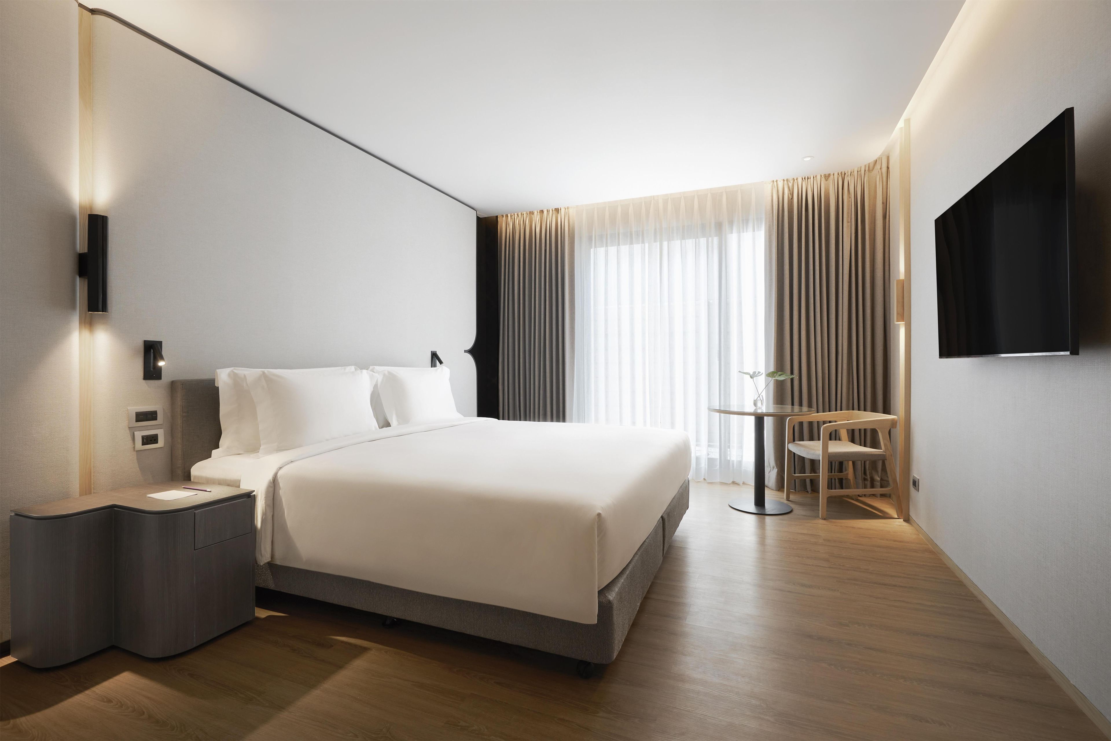A hotel room with a bed, bedside table, lamp, TV, curtains, and a small table with a chair.