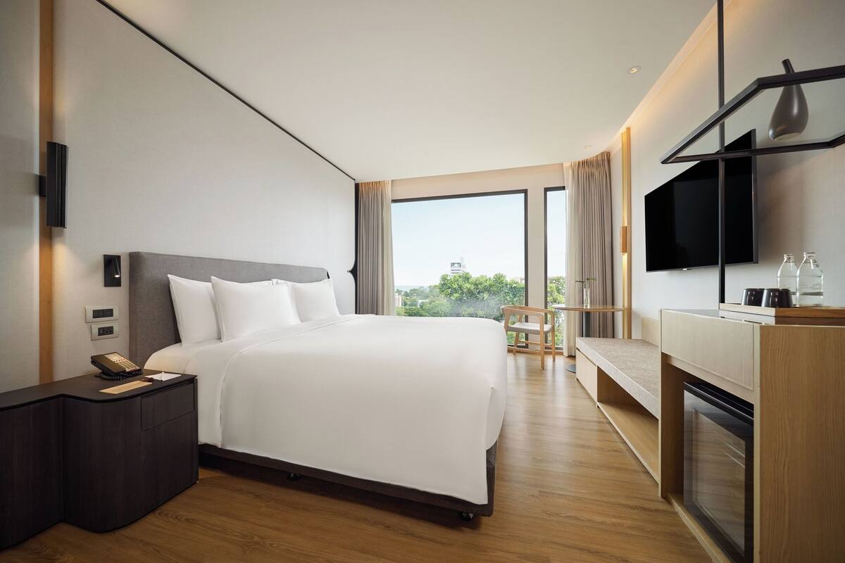 A modern hotel room with a large bed, a flat-screen TV, a bedside table with a phone, and a view of the outdoors.