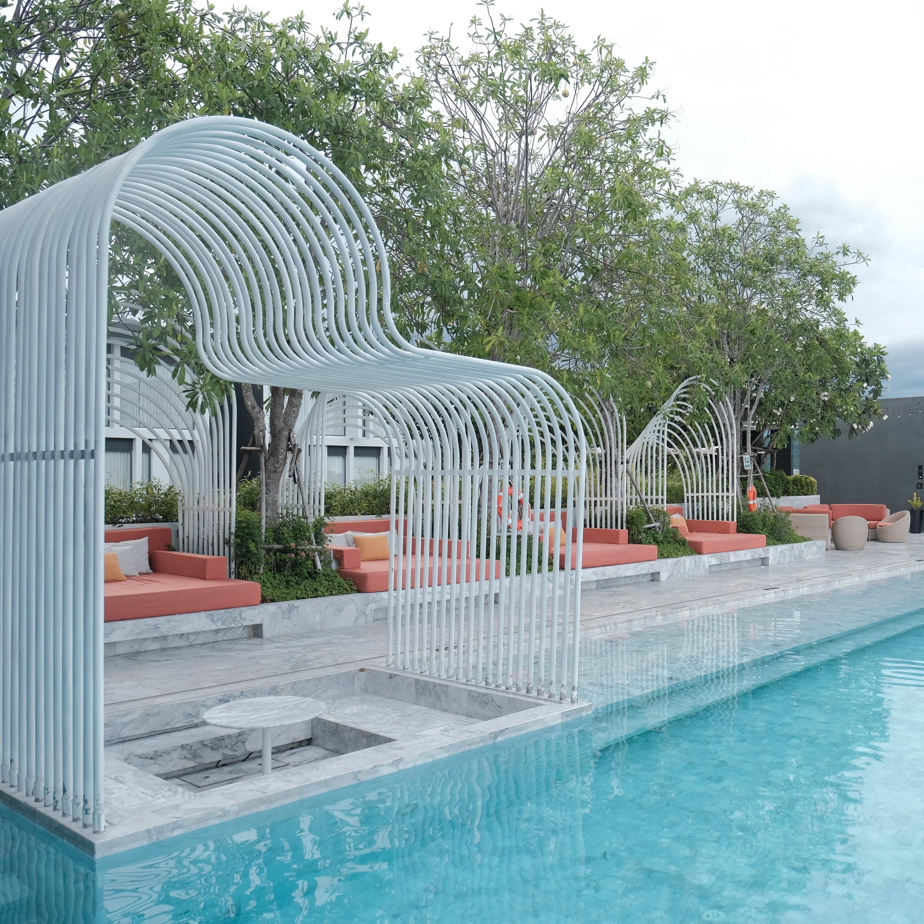 Un espace aménagé au bord de la piscine, avec une structure blanche et incurvée qui offre de l’ombre, un coin salon doté de coussins et une vue dégagée sur la piscine.